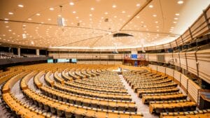 Wide-angle view of the European Parliament hemicycle in Brussels with curved rows of empty wooden desks beneath overhead chamber lighting.