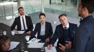 Diverse business executives in suits discuss documents around a table in a glass-walled boardroom