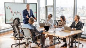 A business leader presents financial charts on a large screen to colleagues seated around a modern boardroom table.