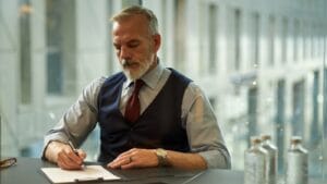 A silver-haired executive in a waistcoat signs a document at a desk in a bright modern office.